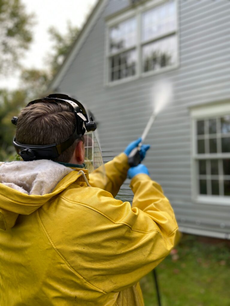 pexels-photo-5652626-5652626 Person cleaning house siding with a pressure washer, wearing protective gear in a yellow raincoat.